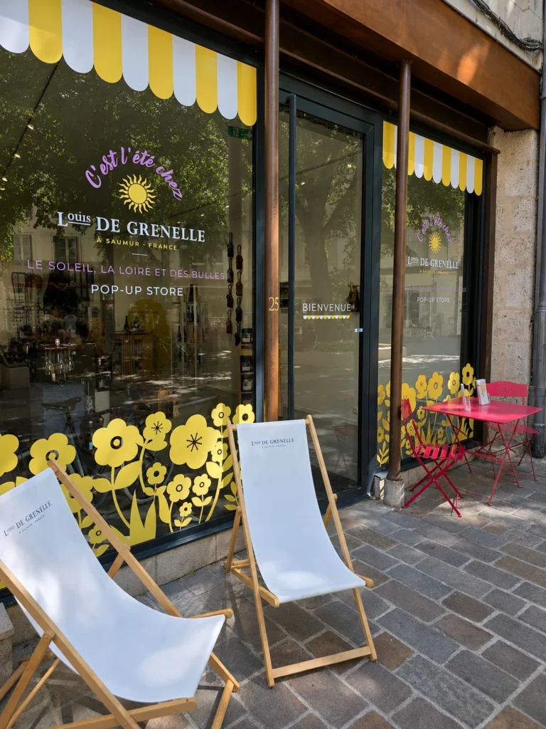 Vitrine de la boutique éphémère Louis de Grenelle à Saumur, décorée d’une vitrophanie sur mesure avec des fleurs jaunes, des motifs estivaux et des lettrages adhésifs personnalisés.