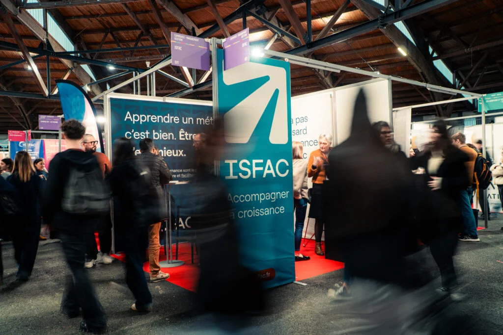 Stand ISFAC lors d’un salon étudiant avec habillage imprimé turquoise, panneaux de signalétique suspendus et visiteurs découvrant les formations proposées.
